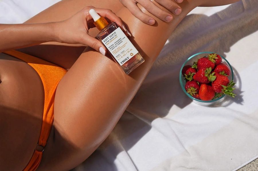 how to treat itchy bikini line | woman in orange bikini on white towel holding glass bottle of Truly's Soft Serve After Shave Oil with bowl of fresh strawberries beside her