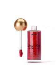 A bottle of Truly Beauty Luxury Peptide Lip Oil with a gold-topped applicator wand stands upright. The shimmering red lip oil is set against a soft white background.