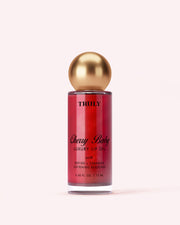 A bottle of Truly Luxury Body Oils by Truly Cherry Baby, with a gold spherical cap and red liquid in a clear container with black text, stands against a pale pink background.