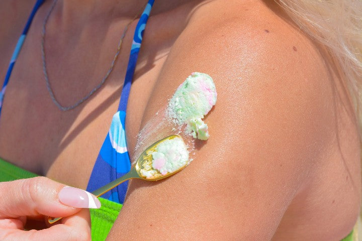 Woman in a blue and green bikini applying Truly's unicorn fruit butter to her shoulder using a gold spoon