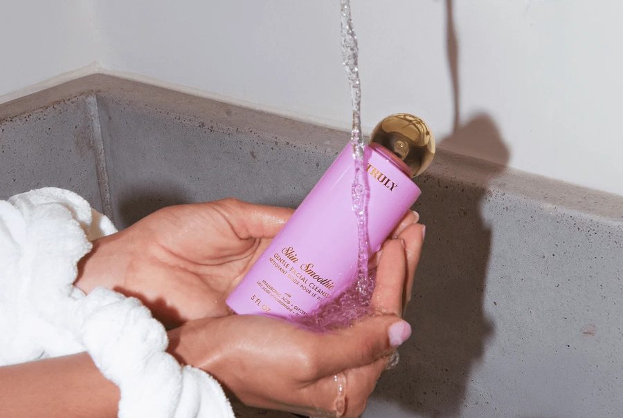 Best gentle cleanser for damaged skin barrier  | A person in a white robe holds a Truly Beauty Skin Smoothie Gentle Facial Cleanser bottle under running water.