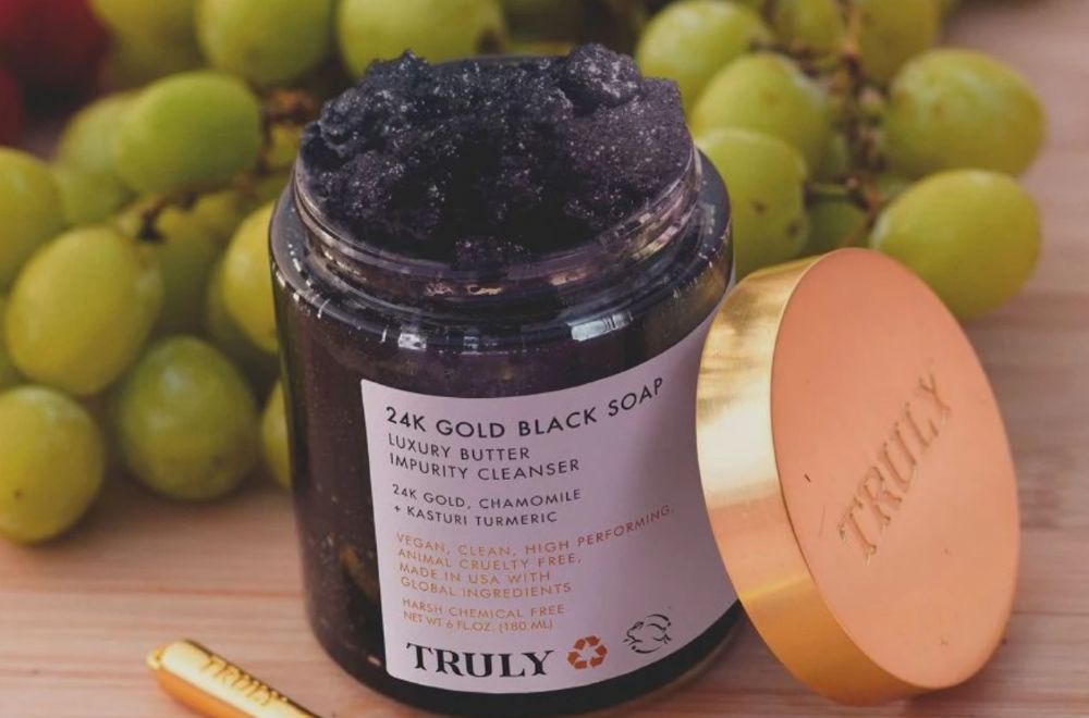 best exfoliating soap | closeup of an open jar of Truly's 24k gold black soap with green grapes in background