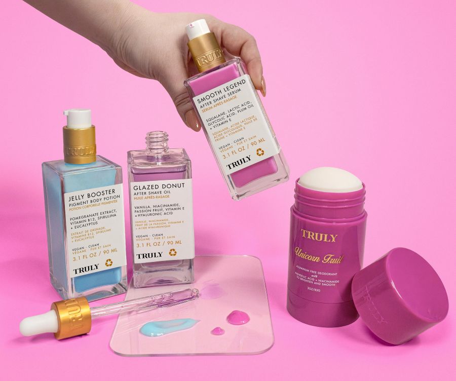 A hand holds a bottle from the Truly Beauty Body Brightening Set above a pink background, with two more serums, an open moisturizer jar, and serum drops for how to get even skin tone on body