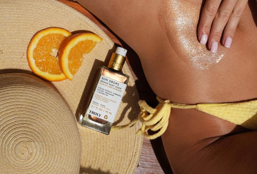 how to self tan instantly | A person in a yellow bikini applies Truly Beauty’s Sun Drops Bronze Body Glow to their torso while lying beside a straw hat, two orange slices, and the product bottle