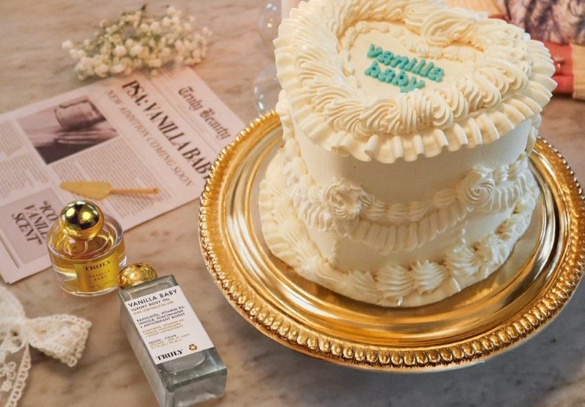 what is a gourmand scent | A vintage-style heart cake with white piped frosting sits on a gold plate alongside glass cosmetic bottles and newspaper