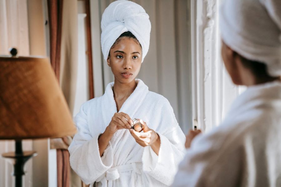 why is my skin so oily | Women in white robe looking in mirror holding jar of face cream
