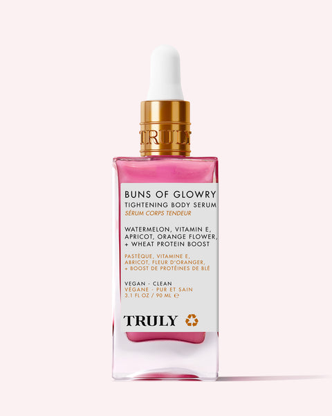 A pink glass bottle of Truly Beauty Buns Of Glowry Tightening Body Serum with a gold dropper. The label highlights key ingredients like watermelon, vitamin E, apricot, and wheat protein for ultra-hydrated, firmer-looking skin.