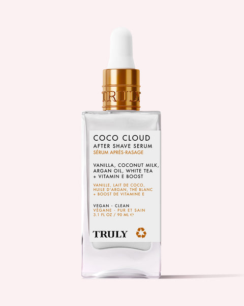 A glass bottle of Truly Beauty’s Coco Cloud After Shave Serum, featuring a white label and gold dropper cap, is displayed against a light pink background.