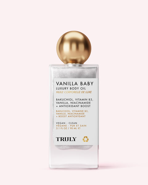 A clear bottle of Truly Beauty’s Vanilla Baby Luxury Body Oil with a gold cap sits on a light pink background; it features anti-aging bakuchiol, vitamin B3, vanilla, and antioxidants.