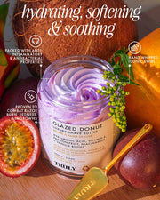 An open jar of Truly Beauty Glazed Donut Luxury Shave Butter with a purple swirl sits among coconuts and passionfruit, showcasing its hydrating, soothing, and anti-inflammatory formula. A gold spatula rests nearby.