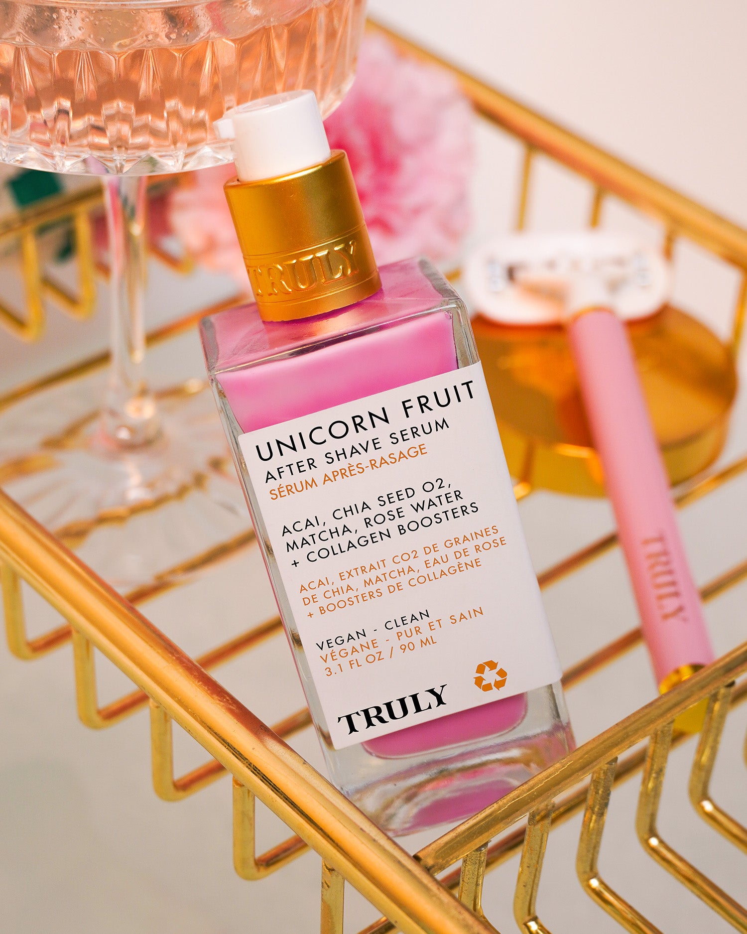 A bottle of Truly Beauty Unicorn Fruit After Shave Serum, loved for soothing post-shave skin and treating ingrown hairs, rests in a gold wire basket beside a pink razor and a glass of rosé wine, with pink flowers softly blurred behind.