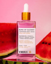 A pink bottle of Truly Beauty’s Buns Of Glowry Tightening Body Serum with a white dropper cap sits among watermelon slices on a pink background. The label displays its key ingredients and product details.