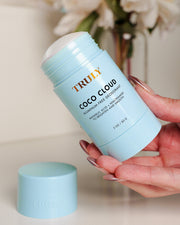 A hand holds a light blue tube of Truly Beauty Coco Cloud Ingrown Prevention and Brightening Deodorant with the cap off, showing the white stick. The cap sits nearby on a beige surface, with blurred flowers in the background.