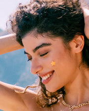 Outdoors, a young woman with curly hair and a gold chain necklace smiles with her eyes closed, sunlight highlighting her face and hair. She wears Truly Beauty's Our Stars Pre-Game Blemish Patch, a yellow star hydrocolloid patch, on her cheek.