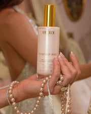 A person holds a bottle of Truly Beauty Diamond Baby Treatment Body Serum with gold text and a metallic cap, alongside a strand of pearls. The softly blurred background adds an elegant touch.