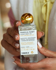 A person with manicured nails holds a bottle of Truly Beauty's Vanilla Baby Luxury Body Oil, featuring a gold cap and white label listing anti-aging ingredients like bakuchiol, niacinamide, vanilla, and antioxidants.