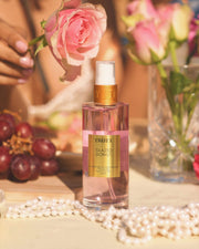A bottle of Truly Beauty Glazed Donut Perfume Mist sits on a table with pearls, grapes, a glass, and pink roses. The sweet scent of whipped buttercream fills the sunny scene as a hand in the background holds one rose.