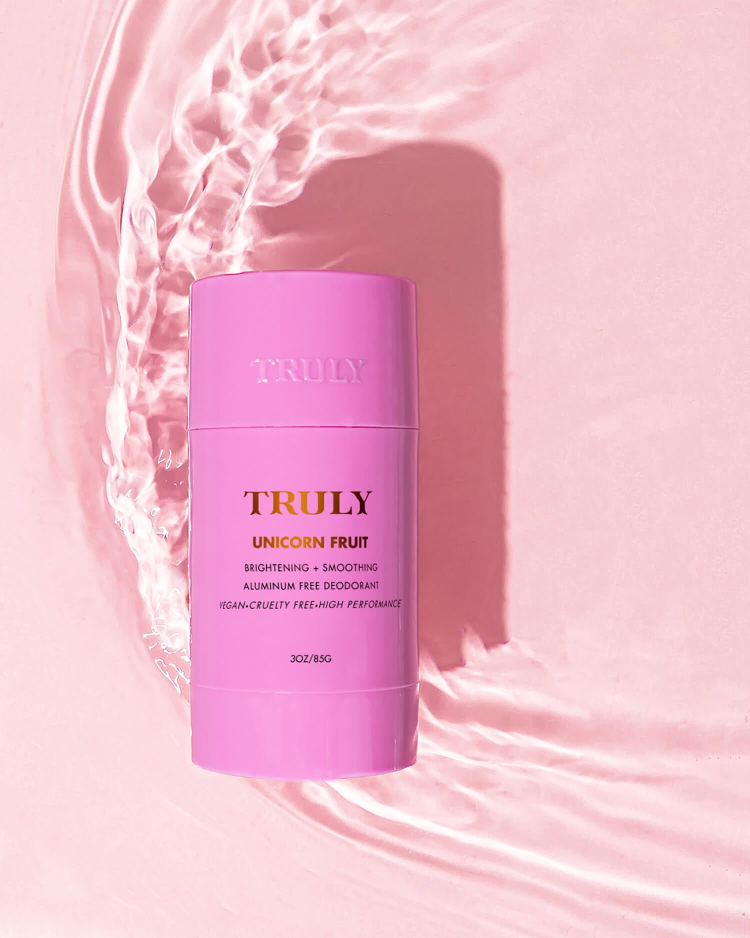 A pink stick from the Truly Beauty Unicorn Fruit Duo stands upright on a shiny, light pink surface with water ripples around it. The label highlights its smoothing, aluminum-free formula and sweet cotton candy scent.