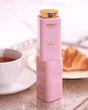 A pink bottle from Truly Beauty's The Body Treatments Complete Collection, celebrated for targeting stretch marks, rests on a tray with tea and a croissant against a soft, bright background.