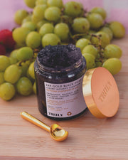 An open jar of Truly Beautys 24K Gold Black Soap Luxury Impurity Cleanser with a gold lid and matching spoon rests on wood, green grapes in the background. This luxurious cleanser delivers exfoliation and anti-aging benefits to your skincare routine.