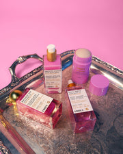 Truly Beauty’s Tiktok Pink Routine—including skin-smoothing creams and shimmering body oil in pink and purple bottles and jars—is displayed on a silver tray with a pink background. Some bottles have droppers; one jar shows cream inside.