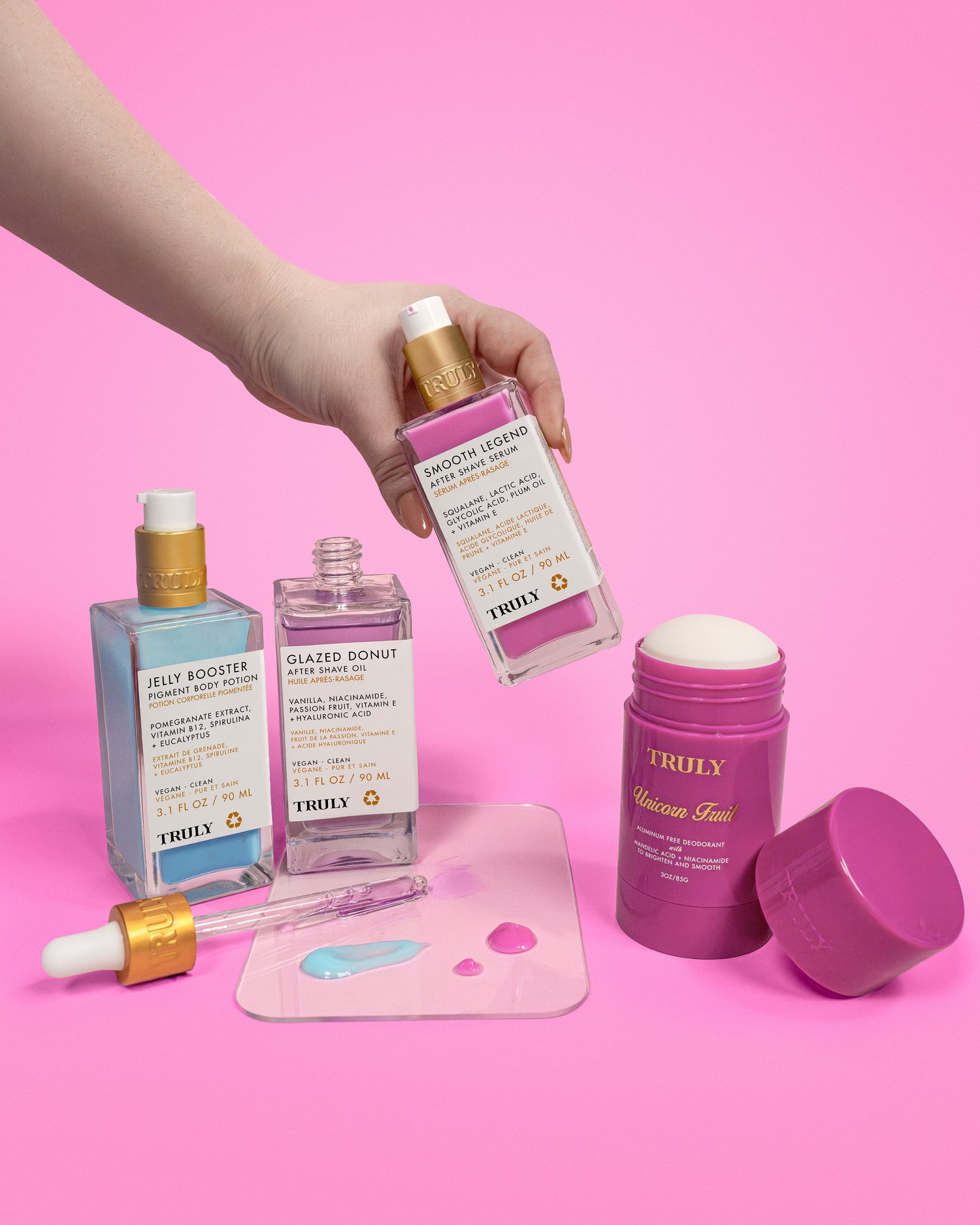 A hand holds a bottle from the Truly Beauty Body Brightening Set above a pink background, with two more serums, an open moisturizer jar, and serum drops—perfect for brightening and evening your skin tone.