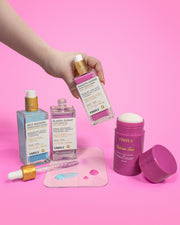 A hand holds a bottle from the Truly Beauty Body Brightening Set above a pink background, with two more serums, an open moisturizer jar, and serum drops—perfect for brightening and evening your skin tone.
