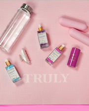 A pink exercise mat featuring “TRULY,” with a clear water bottle, two pink dumbbells, a white hair clip, a purple deodorant stick, and Truly Beauty’s Body Brightening Set for an even skin tone.