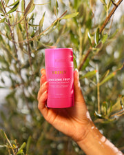 A hand holds a pink container of Truly Beauty’s Unicorn Fruit Skin Brightening and Smoothing Deodorant in front of green leafy branches.