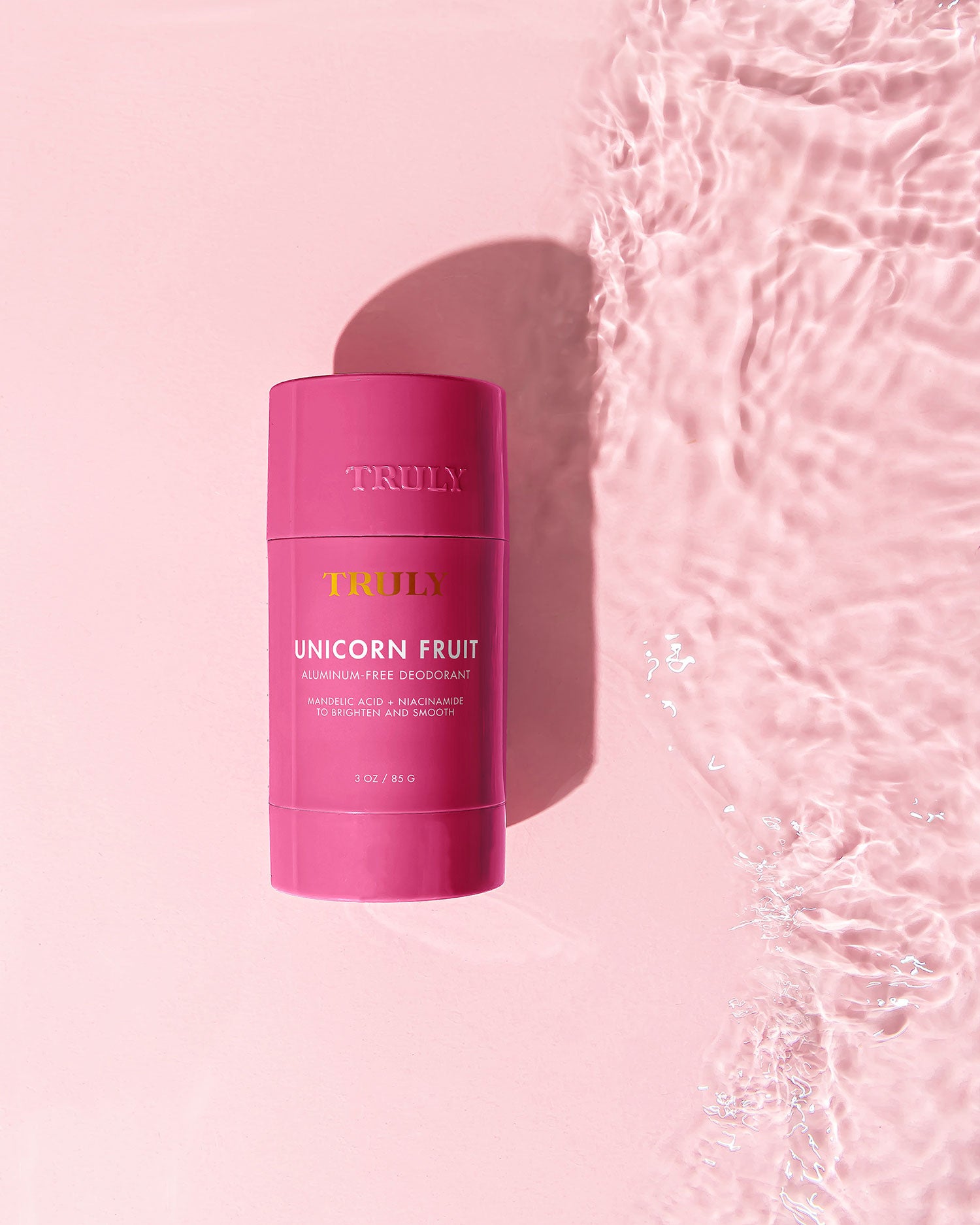 A vibrant stick of Truly Beauty’s Unicorn Fruit Duo deodorant sits on a smooth pink surface beside glittering water, evoking a bright, refreshing vibe with notes of cotton candy.