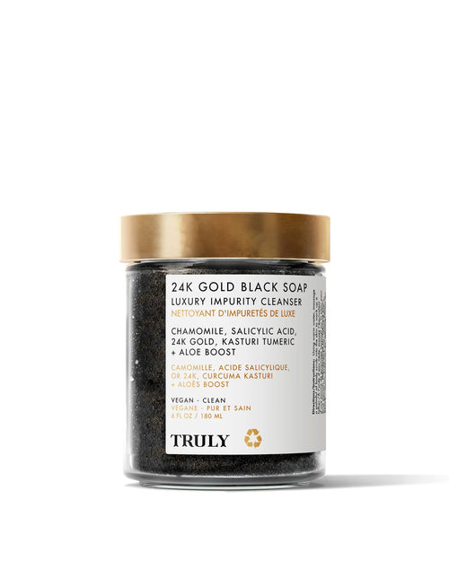 A clear jar with a gold lid labeled 24K Gold Black Soap Luxury Impurity Cleanser by Truly Beauty, showing African black soap inside. The label highlights exfoliating ingredients like chamomile, salicylic acid, gold, and turmeric.