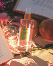 A bottle of Truly Beauty’s Birthday Sex Perfume Mist, featuring a spicy floral scent with jasmine notes, rests on peach fabric amid pearls, flowers, peaches, and open books in warm sunlight.