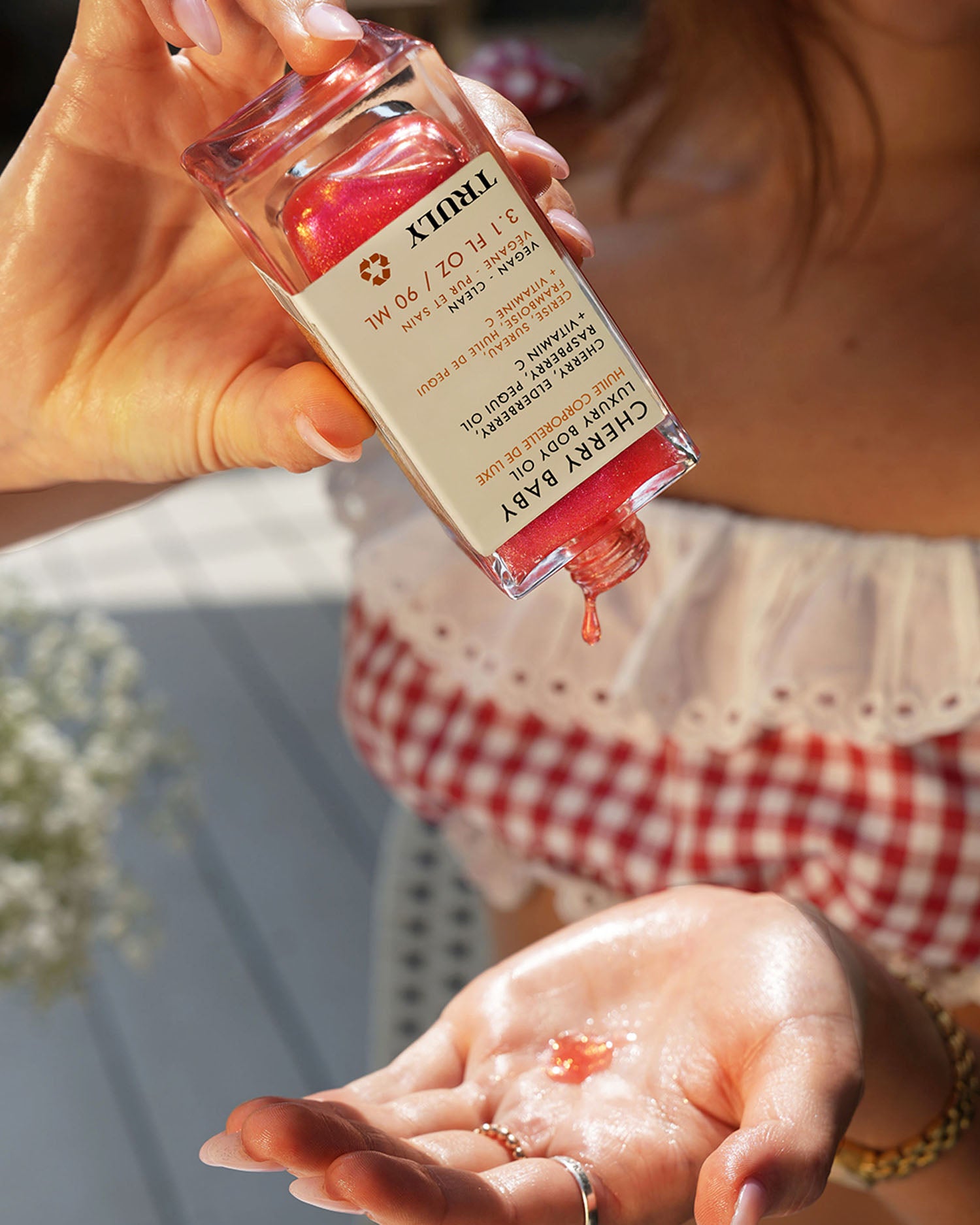 A woman pours Truly Beauty’s Cherry Baby Luxury Body Oil onto her palm, dressed in a red and white checkered top with jewelry as sunlight enhances the glowing, radiant scene.