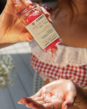 A woman pours Truly Beauty’s Cherry Baby Luxury Body Oil onto her palm, dressed in a red and white checkered top with jewelry as sunlight enhances the glowing, radiant scene.