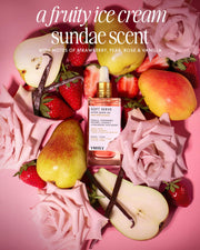 A bottle of Truly Beauty’s Soft Serve After Shave Oil is surrounded by pink roses, strawberries, pears, vanilla beans, and strawberry slices on a pink background, with white text describing a fruity ice cream sundae scent.