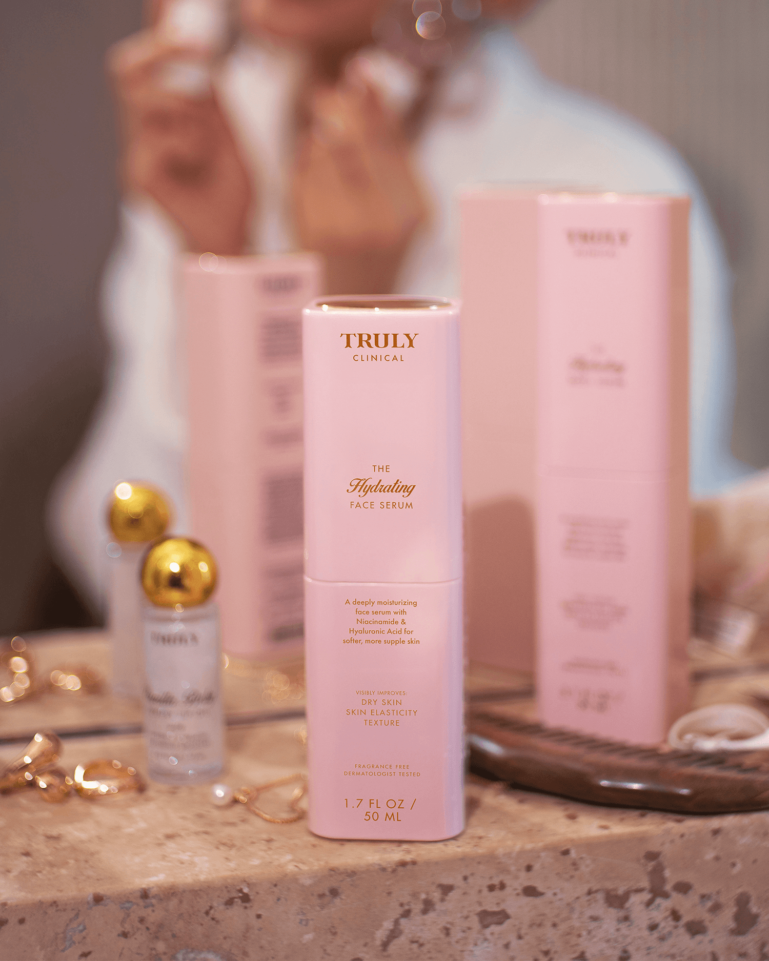 A bottle of Truly Beauty’s The Face Serum Duo rests on a bathroom counter next to its pink packaging, jewelry, and other skincare items, while someone in the background applies this brightening serum.