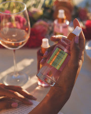 A hand holds a bottle of Truly Beauty Birthday Sex Perfume Mist with a bright label. In the background, a glass of rosé wine, blurred flowers, and other bottles sit on a sunlit table, reflecting its spicy jasmine floral scent.