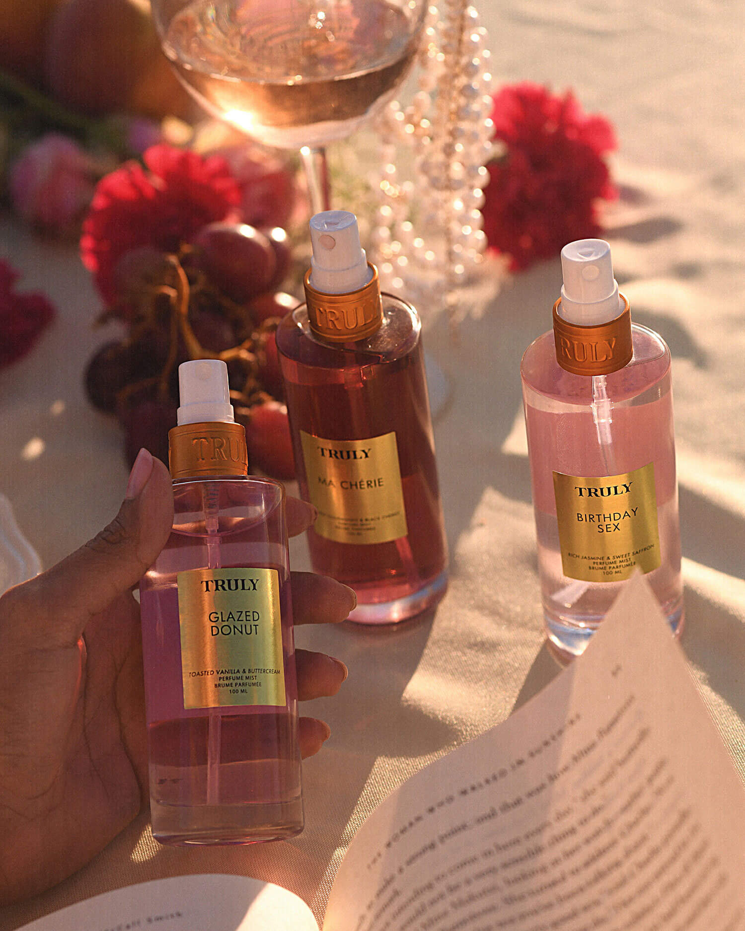 A hand holds a bottle of Truly Beautys Glazed Donut Perfume Mist, surrounded by Unicorn Fruit and Birthday Sex sprays on a sunlit table with flowers, fruit, a wine glass, and an open book.