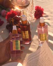 A hand holds a bottle of Truly Beautys Glazed Donut Perfume Mist, surrounded by Unicorn Fruit and Birthday Sex sprays on a sunlit table with flowers, fruit, a wine glass, and an open book.