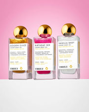 Three Truly Beauty Birthday Sex Luxury Body Oils with gold caps are shown on a pink and white background, with the front label highlighting ingredients such as hyaluronic acid and peptides.
