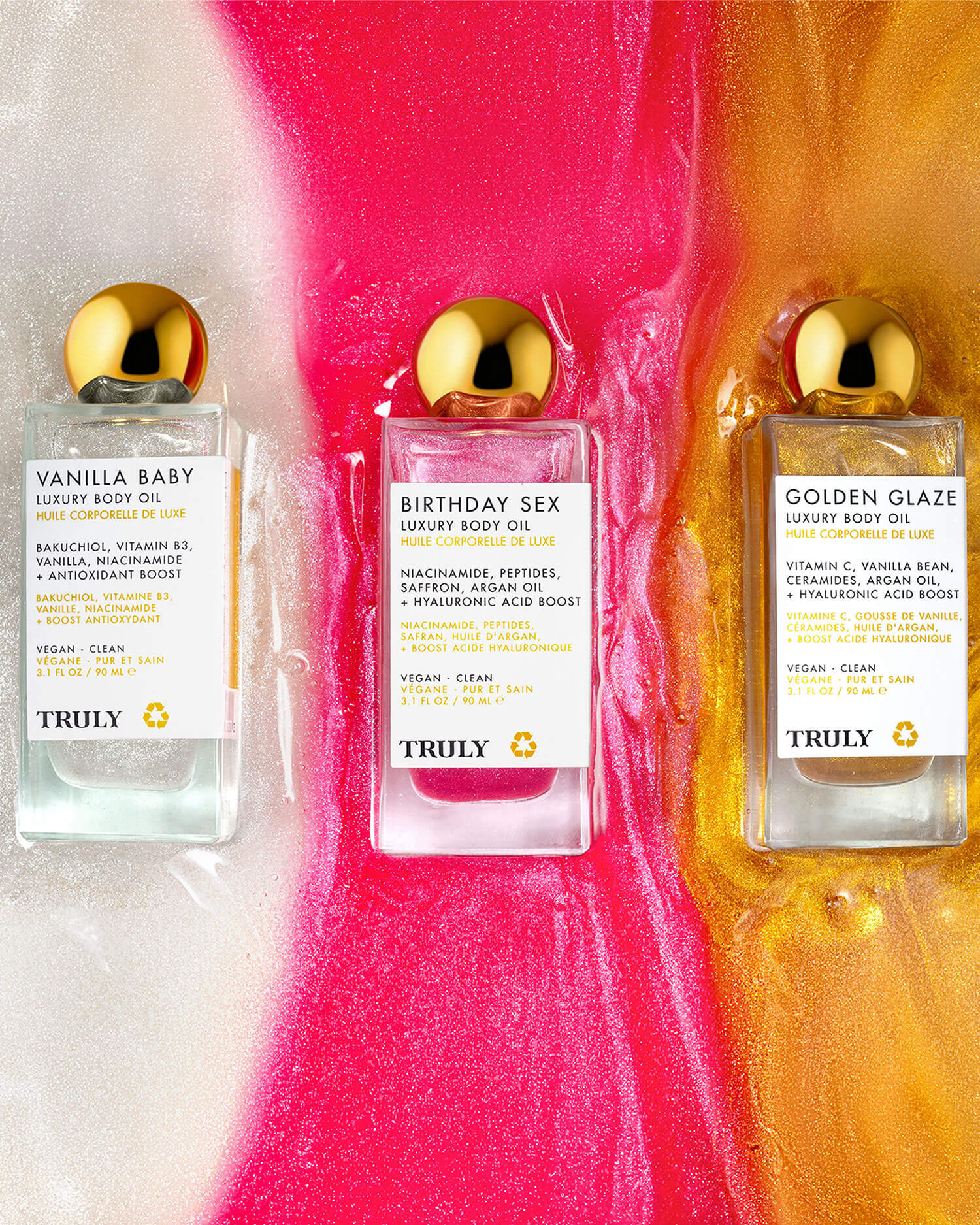 Three bottles of Truly Beauty Birthday Sex Luxury Body Oil are displayed on a glossy surface with pink, white, and gold swirls. Each bottle features a gold cap and white label enriched with peptides.