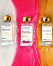 Three bottles of Truly Beauty Birthday Sex Luxury Body Oil are displayed on a glossy surface with pink, white, and gold swirls. Each bottle features a gold cap and white label enriched with peptides.
