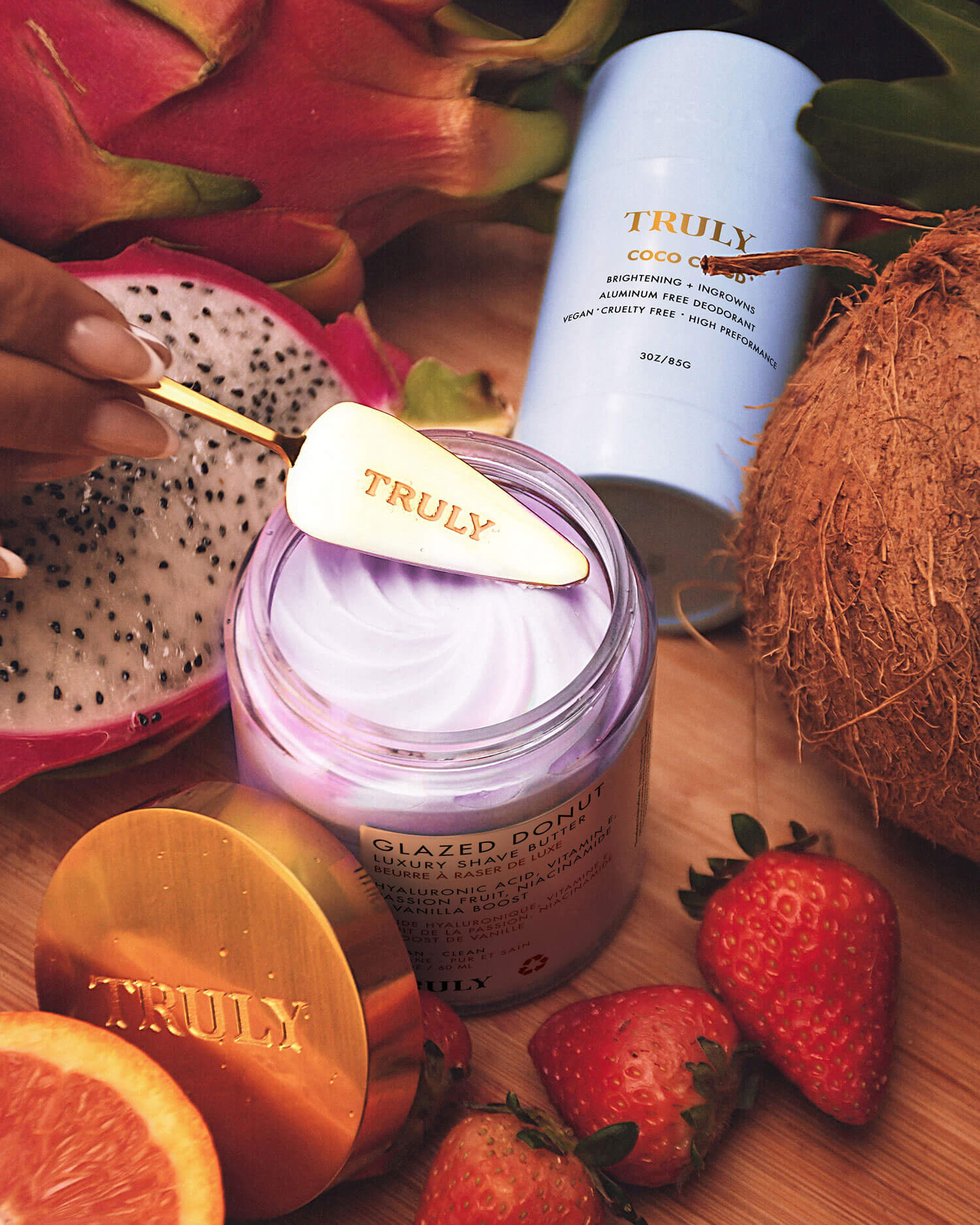 A hand spreads Truly Beauty’s Glazed Donut Luxury Shave Butter with a gold spatula, surrounded by dragon fruit, coconut, strawberries, and a blue bottle labeled Truly Coco Cloud.