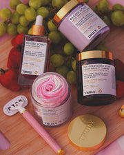 Four Truly Beauty products, including the 24K Gold Black Soap Luxury Impurity Cleanser, jars labeled Glazed Donut, a Soft Serve cream, and an after shave oil, are arranged on a wooden surface with grapes, strawberries, and a pink razor.