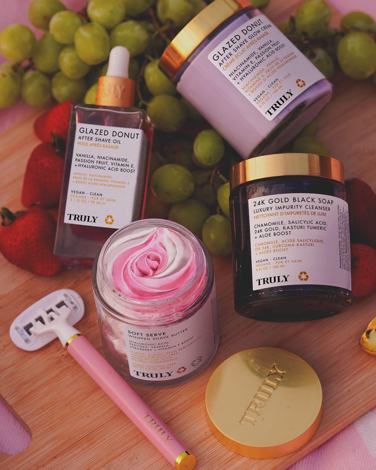 A flat lay features Glazed Donut After Shave Oil by Truly for post-shave hydration, a pink razor, 24K Gold Black Soap, Soft Serve Whipped Body Butter for soft skin, fresh grapes and strawberries, and a gold Truly lid on a wooden surface.