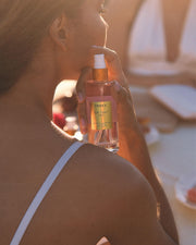 A woman in a white strap tank top holds a pink bottle of Truly Beautys Birthday Sex Perfume Mist with jasmine notes near her shoulder, against a sunlit, softly blurred background.
