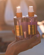 A hand holds two spray bottles—Truly Beauty Birthday Sex and Glazed Donut Perfume Mist—gleaming with gold labels as warm sunlight highlights their decadent whipped buttercream and toasted vanilla fragrance notes.