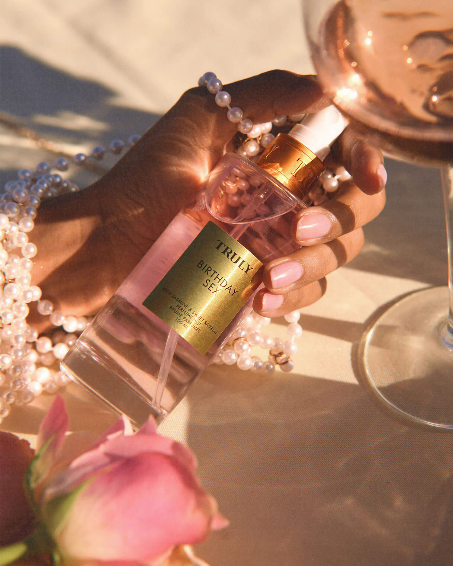 A manicured hand wearing a pearl bracelet holds Truly Beauty’s Birthday Sex Perfume Mist, a spicy floral scent. Nearby, a wine glass and pink rose sit on a sunlit, cream-colored surface.