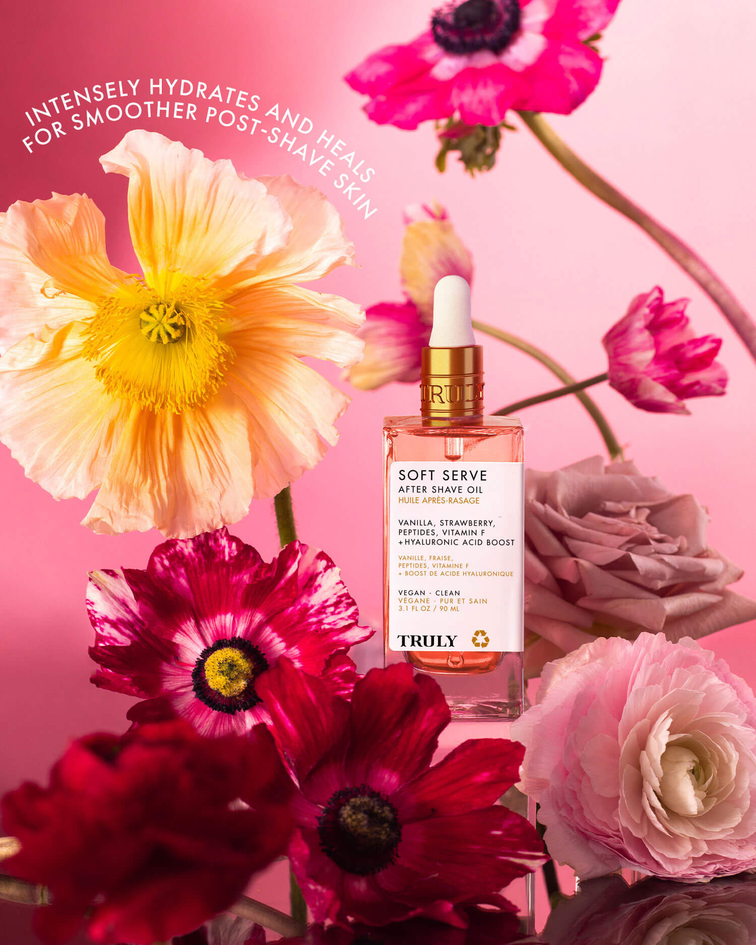 A bottle of Truly Beauty Soft Serve After Shave Oil sits among colorful flowers on a pink background, with white text above: “Intensely hydrates and heals for smoother post-shave skin.”.