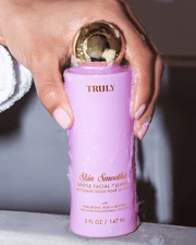 A hand with light pink nails holds a pink bottle of Truly Beauty Skin Smoothie Gentle Facial Cleanser with a gold cap and white and gold text, water running down it. The person is wearing a white robe.